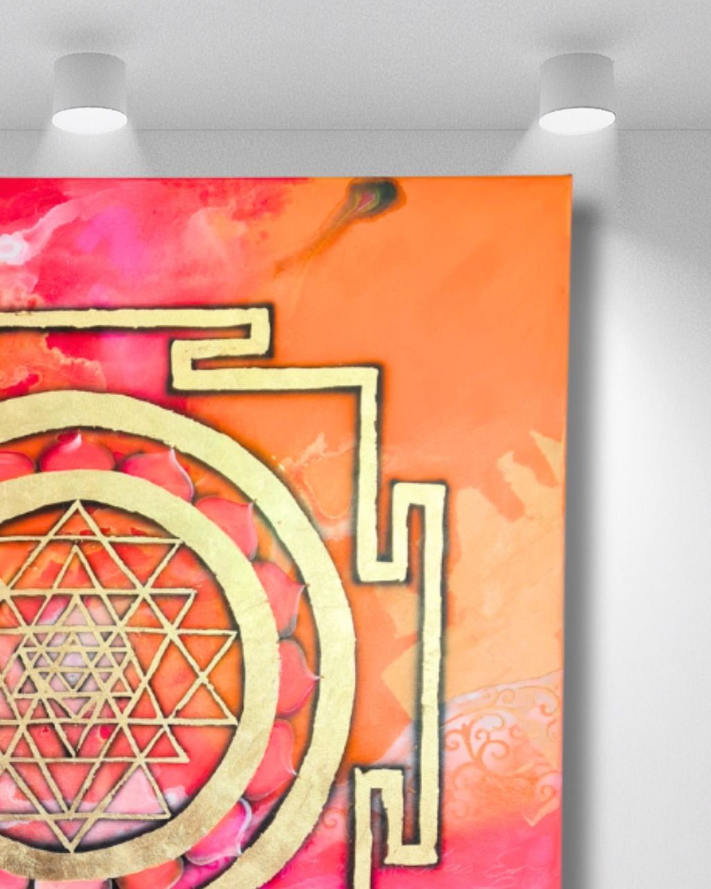 A framed Sri Yantra symbol with intricate geometric patterns in gold and red tones mounted on a white wall under two ceiling lights.